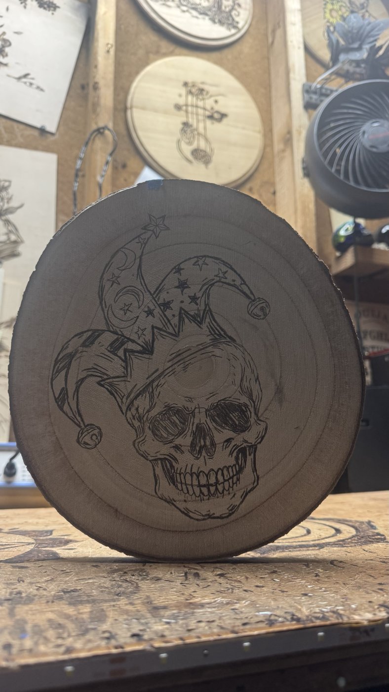 Jester skull with a star-and-moon hat and bells, linework burned into a wood round